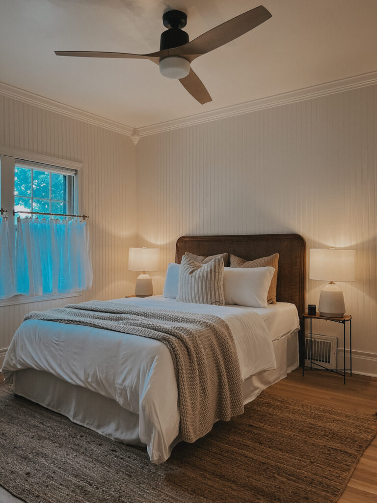 Image of bedroom with white beadboard walls, a modern but classic ceiling fan, simple half curtains, a rattan headboard, and white and neutral bedding.