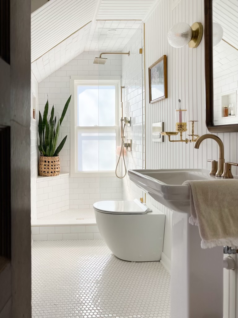White and brass upstairs bathroom. The walls and ceiling are white beadboard, there is a white pedestal sink, the shower surround is white subway tile, and all accents are brass.