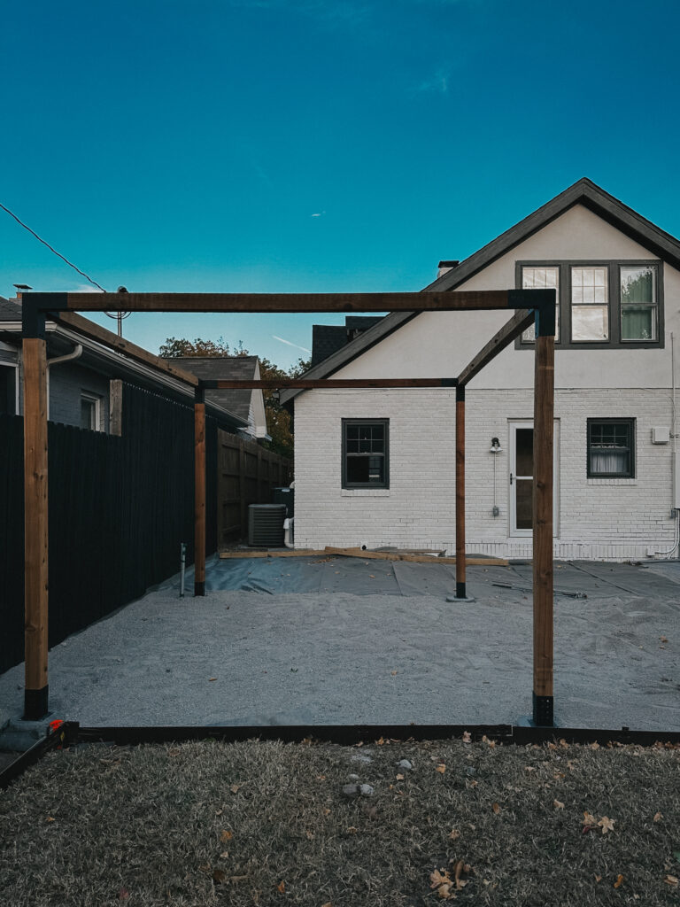 First stage of pergola build completed, with all posts installed and secured to concrete pavers. Gravel covers the ground, and the house is freshly painted.