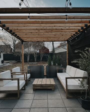 Image of pergola build, strung with lights. Furniture sits inside the pergola space, which was built on a base of gravel and pavers.