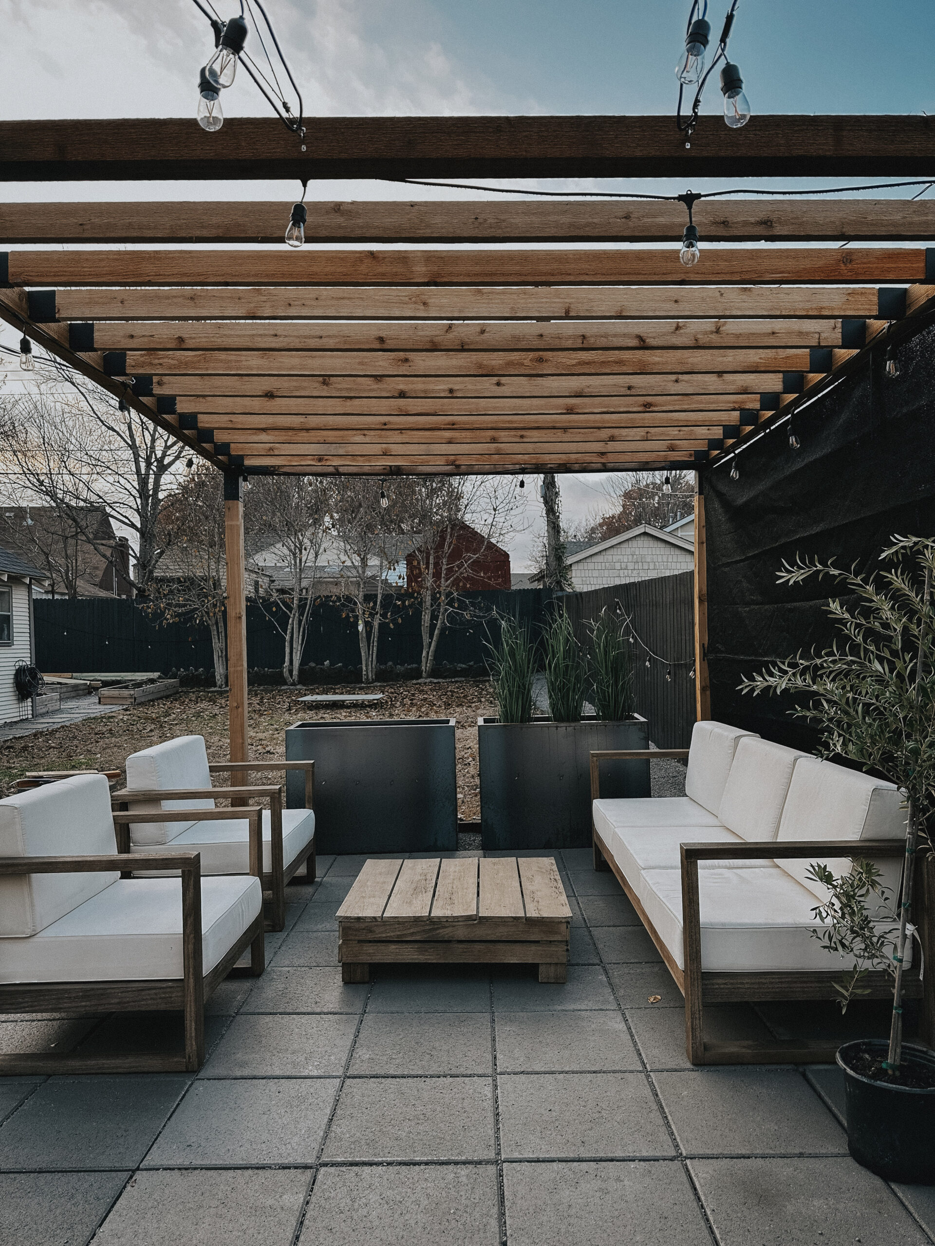 Image of pergola build, strung with lights. Furniture sits inside the pergola space, which was built on a base of gravel and pavers.
