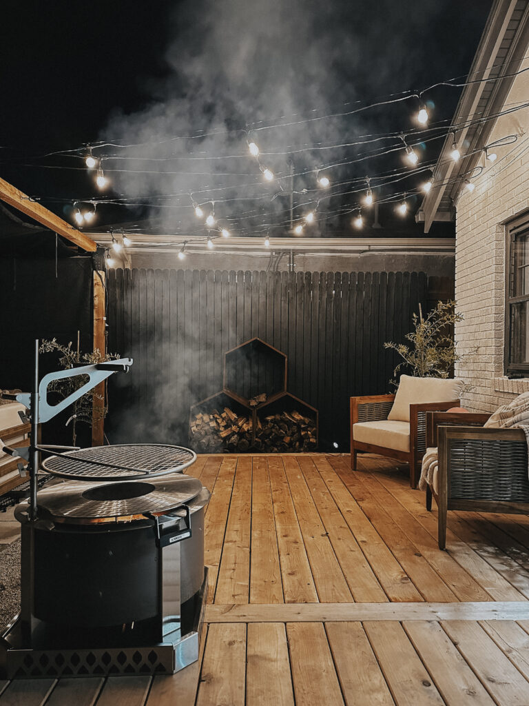 A nighttime scene of deck space, with lights strung overhead, and the firepit smoking.